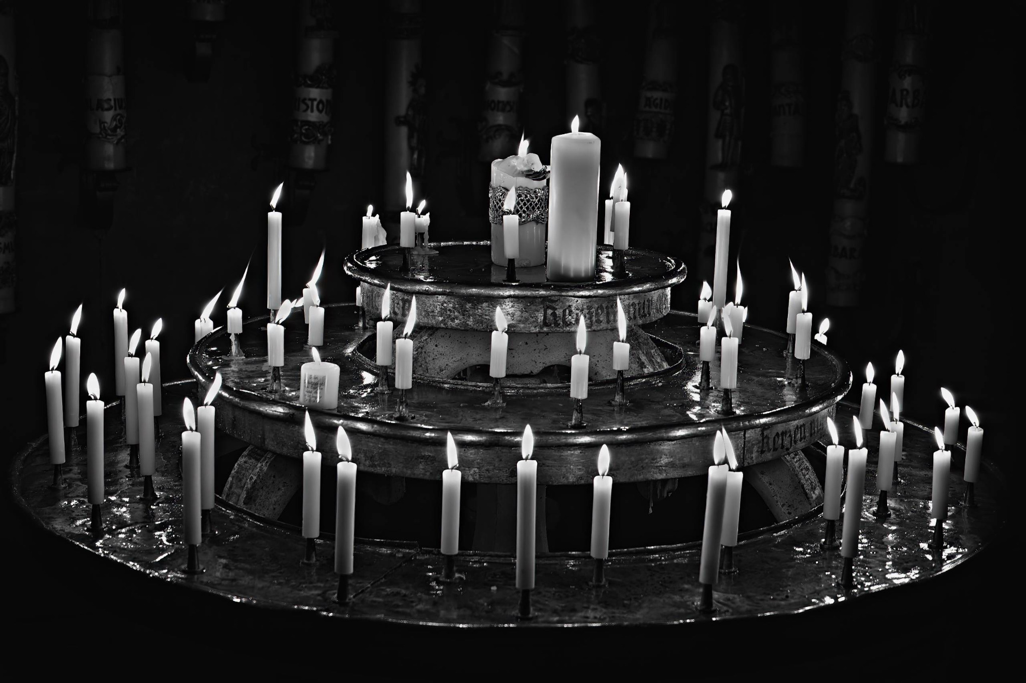 Brennende weiße Votivkerzen stehen auf einem mehrstufigen runden Metallleuchter vor fast schwarzem Hintergrund in einer Kirche in Ebrach.