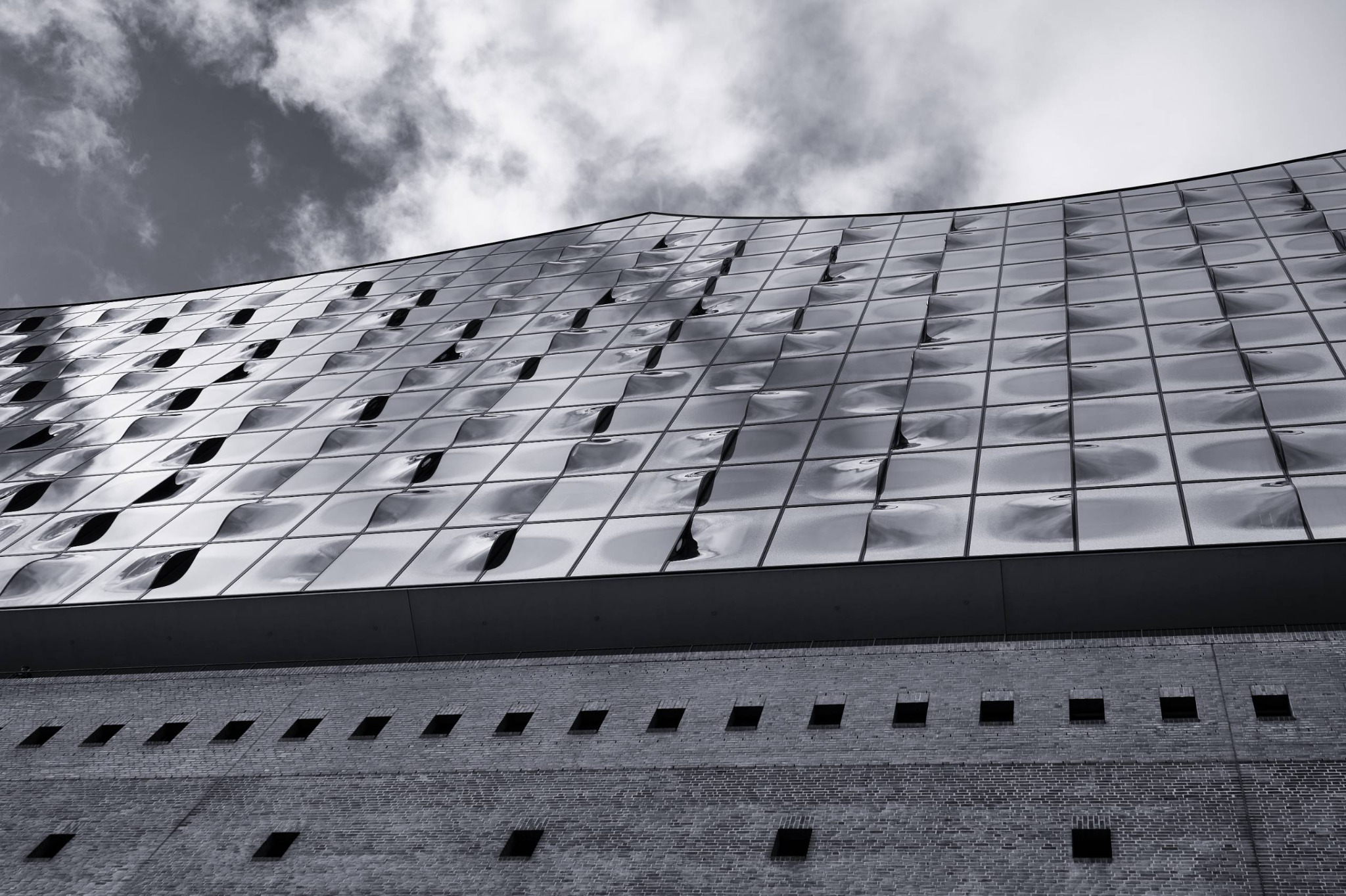 Die spiegelnde Glasfassade der Elbphilharmonie zieht sich diagonal durch das Bild und reflektiert einen wolkigen Himmel über der dunklen Gebäudebasis.