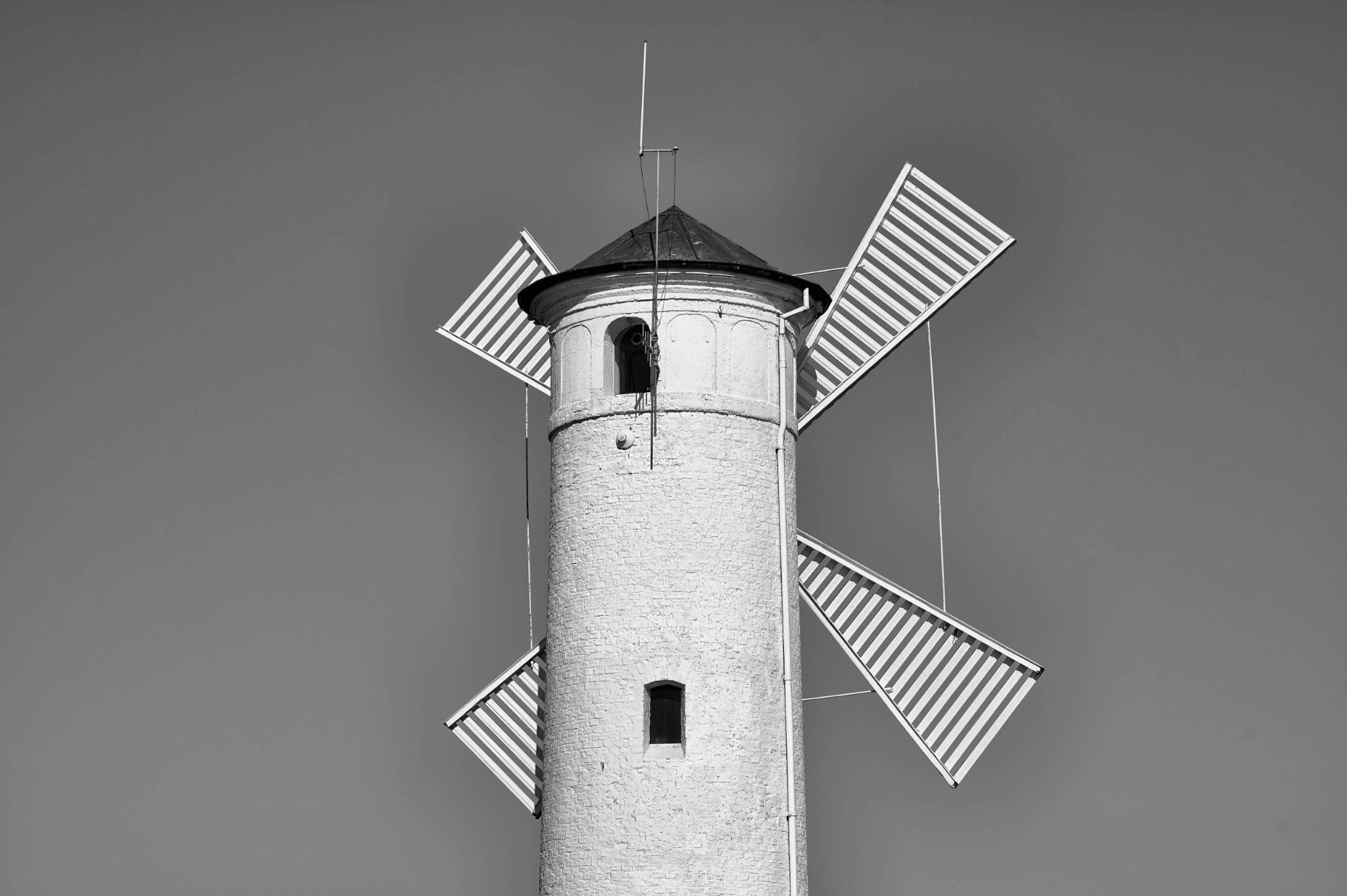 Ein heller Windmühlenturm mit vier gestreiften Flügeln steht freigestellt vor einem gleichmäßig grauen Himmel in Świnoujście.
